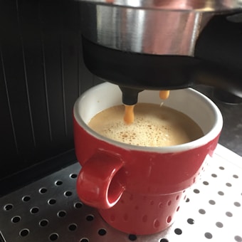 A coffee machine pours espresso into a red mug, which sits on a metal drip tray. The espresso has a light, creamy froth on top.