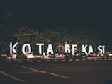 Large illuminated letters spell out 'KOTA BEKASI' along a busy street during the night. Numerous cars are parked or moving alongside the letters, and a few pedestrians are visible. The scene is set against the backdrop of trees and the dark sky.