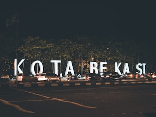 Large illuminated letters spell out 'KOTA BEKASI' along a busy street during the night. Numerous cars are parked or moving alongside the letters, and a few pedestrians are visible. The scene is set against the backdrop of trees and the dark sky.