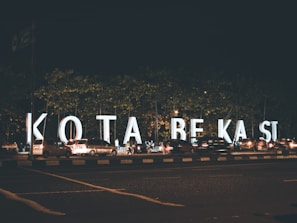 Large illuminated letters spell out 'KOTA BEKASI' along a busy street during the night. Numerous cars are parked or moving alongside the letters, and a few pedestrians are visible. The scene is set against the backdrop of trees and the dark sky.