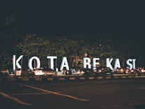 Large illuminated letters spell out 'KOTA BEKASI' along a busy street during the night. Numerous cars are parked or moving alongside the letters, and a few pedestrians are visible. The scene is set against the backdrop of trees and the dark sky.