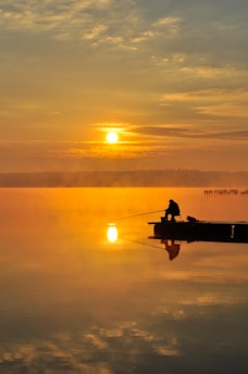 A serene scene of a person fishing on a tranquil lake during sunrise. The sun is rising above the horizon, casting a golden glow across the water and sky. The silhouette of the person sitting on a small boat is reflected on the calm surface of the lake, creating a peaceful and reflective atmosphere.