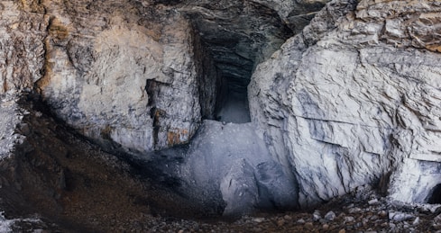 The image captures the interior of a large cave or mine with rocky, uneven surfaces. The walls and ceiling are composed of textured stone with varying shades of gray and brown. The ground is covered in loose rocks and debris.