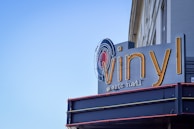 A sign for 'Vinyl Music Hall' is prominently displayed against a building facade. The word 'vinyl' is written in large, yellow, stylized letters with a record graphic integrated into the design. Below, in smaller text, 'music hall' is written in a more subtle font. The sign is fixed to a structure with a dark blue and gray color scheme, contrasting with the clear blue sky in the background.