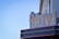 A sign for 'Vinyl Music Hall' is prominently displayed against a building facade. The word 'vinyl' is written in large, yellow, stylized letters with a record graphic integrated into the design. Below, in smaller text, 'music hall' is written in a more subtle font. The sign is fixed to a structure with a dark blue and gray color scheme, contrasting with the clear blue sky in the background.