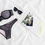 A neatly arranged flat lay featuring a black mesh bikini, dark sunglasses, a passport, a phone with a leaf-patterned case, currency notes, an open planner with marked dates, and a highlighter, all set against a white background.