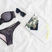 A neatly arranged flat lay featuring a black mesh bikini, dark sunglasses, a passport, a phone with a leaf-patterned case, currency notes, an open planner with marked dates, and a highlighter, all set against a white background.