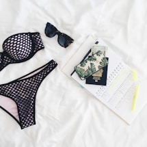 A neatly arranged flat lay featuring a black mesh bikini, dark sunglasses, a passport, a phone with a leaf-patterned case, currency notes, an open planner with marked dates, and a highlighter, all set against a white background.