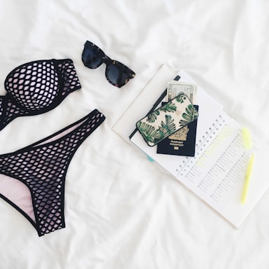 A neatly arranged flat lay featuring a black mesh bikini, dark sunglasses, a passport, a phone with a leaf-patterned case, currency notes, an open planner with marked dates, and a highlighter, all set against a white background.