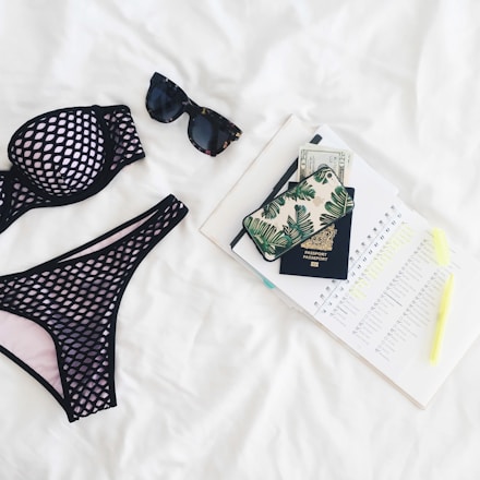 A neatly arranged flat lay featuring a black mesh bikini, dark sunglasses, a passport, a phone with a leaf-patterned case, currency notes, an open planner with marked dates, and a highlighter, all set against a white background.