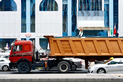 A large red truck with a brown container trailer is parked on the road in front of a modern building with blue-tinted windows. Multiple cars are visible around the truck, and several flags are seen at the entrance of the building. The building has the signage of a cafe and other establishments.