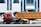 A large red truck with a brown container trailer is parked on the road in front of a modern building with blue-tinted windows. Multiple cars are visible around the truck, and several flags are seen at the entrance of the building. The building has the signage of a cafe and other establishments.