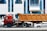A large red truck with a brown container trailer is parked on the road in front of a modern building with blue-tinted windows. Multiple cars are visible around the truck, and several flags are seen at the entrance of the building. The building has the signage of a cafe and other establishments.