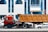 A large red truck with a brown container trailer is parked on the road in front of a modern building with blue-tinted windows. Multiple cars are visible around the truck, and several flags are seen at the entrance of the building. The building has the signage of a cafe and other establishments.