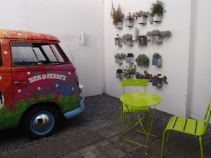 A colorful vintage van with a psychedelic design and the Ben & Jerry's logo is parked in a small courtyard. Next to the van, there is a bright green metal table and chair set placed on a cobblestone surface. On the white wall, a vertical garden of small potted plants is arranged in a neat grid.