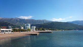 A coastal landscape with a clear blue sky, mountains in the background, and a body of water in the foreground. There are buildings, including high-rise and commercial structures, along the shoreline with some greenery and trees. The water appears calm and there is a pier extending into the water.
