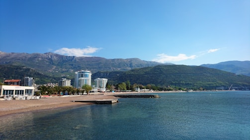 A coastal landscape with a clear blue sky, mountains in the background, and a body of water in the foreground. There are buildings, including high-rise and commercial structures, along the shoreline with some greenery and trees. The water appears calm and there is a pier extending into the water.