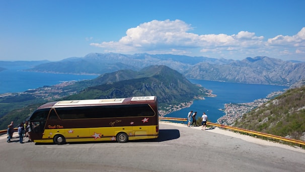 A scenic view from a high vantage point with a large tour bus parked on a road. Several people are standing near the edge, overlooking a breathtaking bay surrounded by mountains. The landscape includes expansive views of water and distant mountains under a clear blue sky with some scattered clouds.