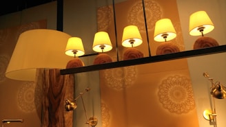 Five warm-colored lamps with beige shades are symmetrically arranged on a dark metal bar. The background features intricate, circular mandala-like patterns in soft tones. A large, elegant floor lamp with a wooden base stands to the left, and several metallic adjustable lights are mounted on the wall.