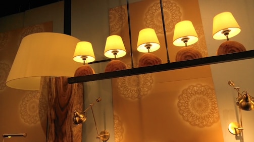 Five warm-colored lamps with beige shades are symmetrically arranged on a dark metal bar. The background features intricate, circular mandala-like patterns in soft tones. A large, elegant floor lamp with a wooden base stands to the left, and several metallic adjustable lights are mounted on the wall.