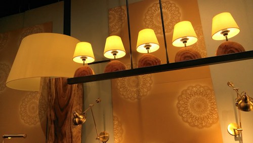 Five warm-colored lamps with beige shades are symmetrically arranged on a dark metal bar. The background features intricate, circular mandala-like patterns in soft tones. A large, elegant floor lamp with a wooden base stands to the left, and several metallic adjustable lights are mounted on the wall.