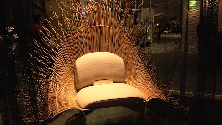 An intricately designed chair with a wicker back resembling waves or a sunburst, set in a dimly lit room. The chair features a soft, cushioned seat with a plaque at the bottom reading 'LOON INTERNATIONAL.' Ambient lighting highlights the natural texture of the wicker material, creating a warm and inviting atmosphere.