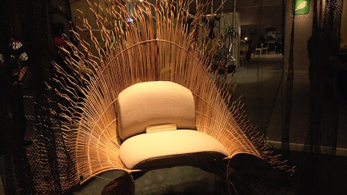 An intricately designed chair with a wicker back resembling waves or a sunburst, set in a dimly lit room. The chair features a soft, cushioned seat with a plaque at the bottom reading 'LOON INTERNATIONAL.' Ambient lighting highlights the natural texture of the wicker material, creating a warm and inviting atmosphere.