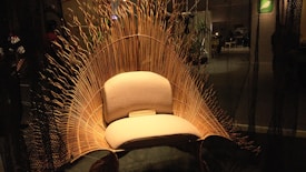 An intricately designed chair with a wicker back resembling waves or a sunburst, set in a dimly lit room. The chair features a soft, cushioned seat with a plaque at the bottom reading 'LOON INTERNATIONAL.' Ambient lighting highlights the natural texture of the wicker material, creating a warm and inviting atmosphere.