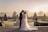 A couple stands in an elegant garden setting, sharing a kiss. The bride wears a flowing white gown with intricate lace details, and the groom is dressed in a dark suit. The garden features manicured hedges and ornamental urns, with a soft, golden sunrise casting a warm glow over the scene and creating a serene atmosphere.