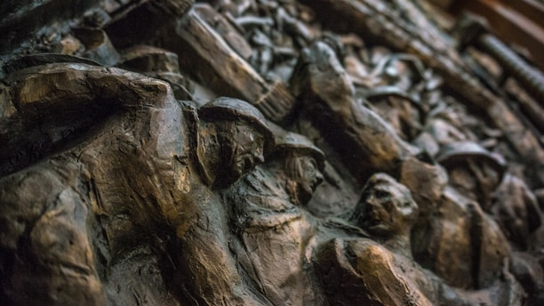 An intricately carved relief featuring several human figures with a dynamic composition, likely depicting a historical or artistic scene. The material appears to be a dark stone or bronze, with shadows accentuating the depth and details of the carving.