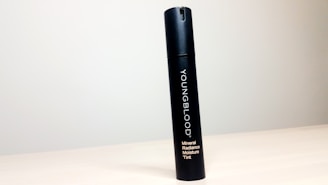 A black cylindrical bottle labeled 'YOUNGBLOOD Mineral Radiance Moisture Tint' stands upright on a light-colored, smooth surface against a plain, light gray background.