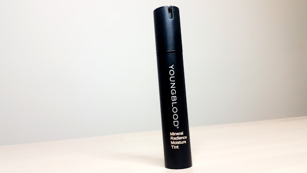 A black cylindrical bottle labeled 'YOUNGBLOOD Mineral Radiance Moisture Tint' stands upright on a light-colored, smooth surface against a plain, light gray background.