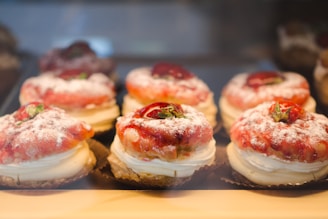 A display of pastries arranged neatly in rows, each topped with powdered sugar, cream, strawberries, and pastry crust. The desserts are placed in a well-lit setting with a warm and inviting atmosphere.
