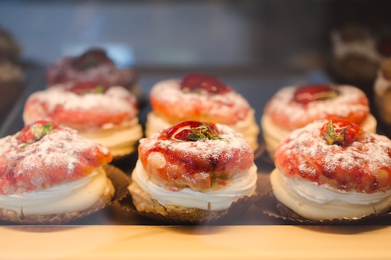 A display of pastries arranged neatly in rows, each topped with powdered sugar, cream, strawberries, and pastry crust. The desserts are placed in a well-lit setting with a warm and inviting atmosphere.