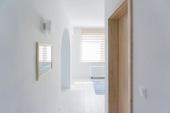 A minimalist hallway with white walls, a wooden door on the right, and a framed picture on the left wall. There is an arched doorway leading to a bright room with large blinds covering a window and a blue striped rug on the floor. The ambiance is clean and spacious.