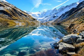 A stunning mountainous landscape reflected in a crystal-clear lake. Snow-covered peaks rise majestically in the background, while rocky terrain and patches of grass and vegetation line the shores. The vibrant blue sky and scattered clouds enhance the scene's natural beauty.