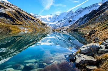 A stunning mountainous landscape reflected in a crystal-clear lake. Snow-covered peaks rise majestically in the background, while rocky terrain and patches of grass and vegetation line the shores. The vibrant blue sky and scattered clouds enhance the scene's natural beauty.