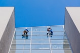 Two workers in protective gear are cleaning the large glass windows of a modern building. They are suspended mid-air by ropes and harnesses, equipped with cleaning tools.