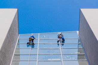 Two workers in protective gear are cleaning the large glass windows of a modern building. They are suspended mid-air by ropes and harnesses, equipped with cleaning tools.