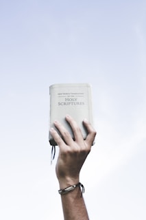 A hand is raised against a clear blue sky, holding up a book titled 'New World Translation of the Holy Scriptures'. The hand is adorned with a simple wristband.