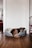 A brown dog with white markings is curled up in a gray and white pet bed. The setting is a room with wooden flooring, white walls, and a visible red chair to the right. In the background, there is a glimpse of another room with a red sofa and a window.