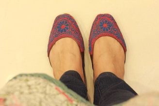 A pair of hands are wearing intricately designed slippers with a floral pattern in red and blue colors. The background is a plain light color, and the person is wearing dark-colored jeans.