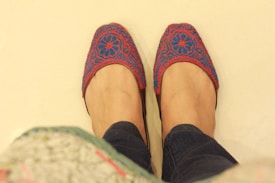 A pair of hands are wearing intricately designed slippers with a floral pattern in red and blue colors. The background is a plain light color, and the person is wearing dark-colored jeans.