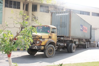 An old, rusty yellow truck with a container trailer attached is parked outside a large industrial building. A small tree with green leaves is positioned towards the left. The industrial building has large windows and a beige exterior.