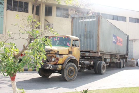An old, rusty yellow truck with a container trailer attached is parked outside a large industrial building. A small tree with green leaves is positioned towards the left. The industrial building has large windows and a beige exterior.