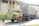 An old, rusty yellow truck with a container trailer attached is parked outside a large industrial building. A small tree with green leaves is positioned towards the left. The industrial building has large windows and a beige exterior.