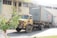 An old, rusty yellow truck with a container trailer attached is parked outside a large industrial building. A small tree with green leaves is positioned towards the left. The industrial building has large windows and a beige exterior.