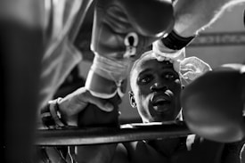 A boxer sits in focus with his mouth slightly open, while a team member wearing gloves tends to his face during a break in a boxing match. The scene is intense and dynamic, captured in black and white, with out-of-focus shadows framing the central figure.