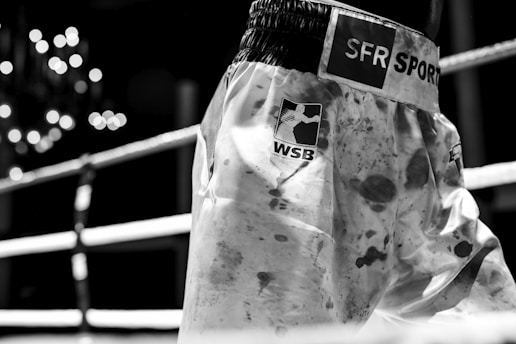A pair of boxing shorts with visible logos and patterns, standing in the foreground with blurred ring ropes and lights in the background.