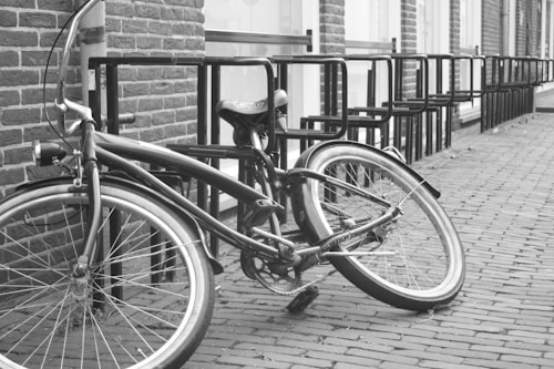 A vintage-style bicycle rests against a brick wall next to a row of empty bike racks. The scene is captured in black and white, highlighting the textures of the cobblestone pavement and the brick wall.